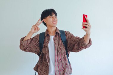 Sırt çantası takmış gülümseyen Asyalı erkek öğrencinin portresi, beyaz arka planda izole edilmiş akıllı telefonuyla selfie çekiyor.