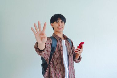 Sırt çantalı gülümseyen Asyalı erkek öğrenci dört parmağı ve telefonu gösteriyor, beyaz arka planda izole edilmiş, üniversite yaşam tarzı konsepti..