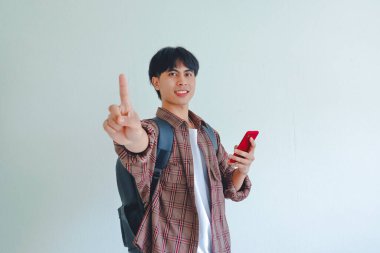 Sırt çantalı, gülümseyen Asyalı kolej çocuğu telefonu tutuyor ve arka planda bir parmağını kameraya doğrultuyor..