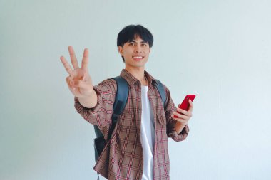Sırt çantalı gülümseyen Asyalı erkek öğrenci, beyaz arka planda izole edilmiş cep telefonunu tutarken kameraya üç parmak işareti gösteriyor..