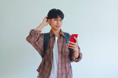 Kafasını kaşıyan sırt çantası taşıyan, kafası karışmış bir cep telefonu tutan bir üniversite öğrencisi.