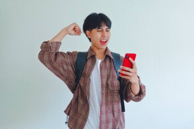 Sırt çantası taşıyan bir üniversite öğrencisi cep telefonunu tutarken mutlu bir ifadeyle başarıyı kutluyor.