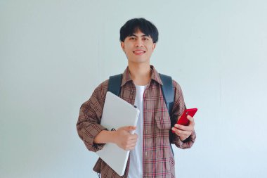 Sırt çantası takmış, elinde dizüstü bilgisayarla gülümseyen Asyalı üniversite öğrencisi beyaz arka plandaki kameraya bakıyor..