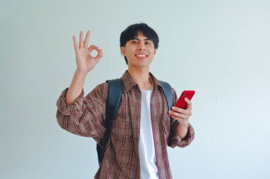 Sırt çantasındaki mutlu Asyalı erkek öğrenci telefonu tutarken el işareti yapıyor. Güvenle gülümsüyor, arka planda izole bir şekilde..
