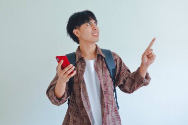 Sırt çantası taşıyan bir erkek üniversite öğrencisi, elinde gülümseyen bir ifadeyle cep telefonu tutarken yanındaki boşluğu işaret ediyor..