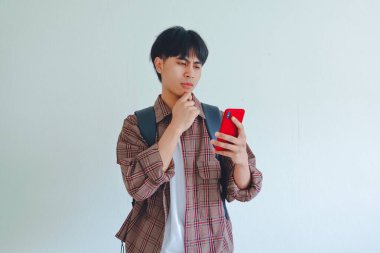 Sırt çantası taşıyan, çenesini tutan ve elinde cep telefonu tutarken düşünen bir üniversite öğrencisi.