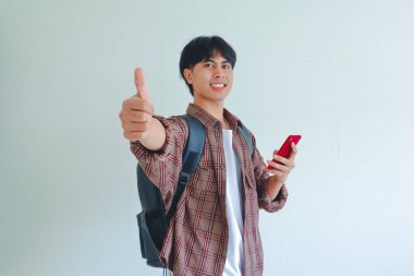 Sırt çantalı, gülümseyen üniversite öğrencisi elinde cep telefonuyla beyaz arka planda izole bir şekilde baş parmak hareketi yapıyor..