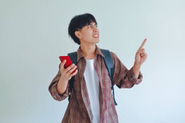 Sırt çantası taşıyan bir erkek üniversite öğrencisi, elinde gülümseyen bir ifadeyle cep telefonu tutarken yanındaki boşluğu işaret ediyor..