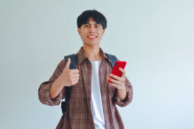 Sırt çantalı, gülümseyen üniversite öğrencisi elinde cep telefonuyla beyaz arka planda izole bir şekilde baş parmak hareketi yapıyor..