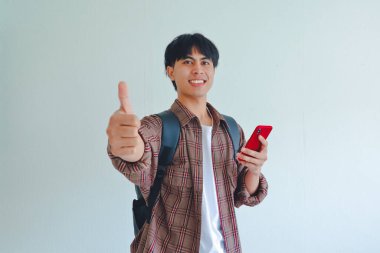 Sırt çantalı, gülümseyen üniversite öğrencisi elinde cep telefonuyla beyaz arka planda izole bir şekilde baş parmak hareketi yapıyor..