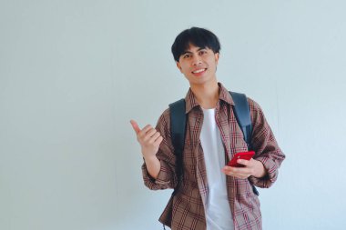 Sırt çantasıyla gülümseyen Asyalı erkek öğrenci elinde telefon tutarken onu işaret ediyor, reklam veya tanıtım konsepti için beyaz üzerine izole edilmiş.