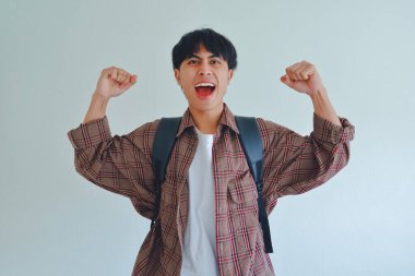 Sırt çantası takmış, yumruklarını iki eliyle sıkan erkek bir üniversite öğrencisi beyaz bir arka planda başarıyı kutlayan mutlu bir ifade sergiliyor.