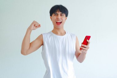 Mutlu Asyalı genç adamın portresi cep telefonu tutan zafer kutlamaları için yumruklarını sıkan beyaz arka planda izole edilmiş zafer ve başarı.