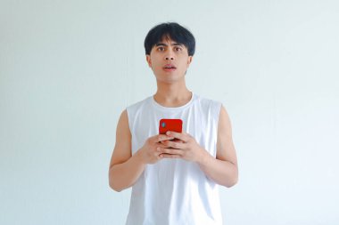 Güneydoğu Asyalı genç bir adamın portresi şok olmuş bir ifadeyle cep telefonu taşıyor, beyaz arka planda izole edilmiş, şaşırmış ve inançsız.