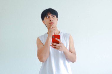 Elinde cep telefonu olan, ciddi bir Asyalı genç adamın portresi beyaz arka planda izole edilmiş, düşünceli bir hareketle yukarı bakıyor.