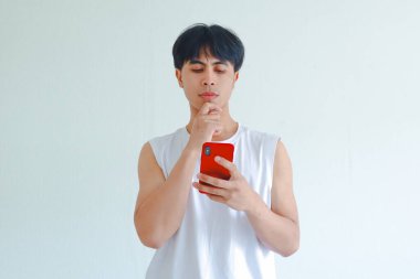 Beyaz arka planda izole edilmiş düşünceli bir ifadeyle cep telefonu tutan Asyalı ciddi bir gencin portresi.
