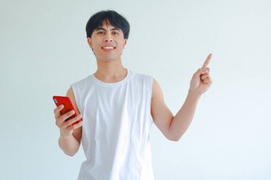 Gülümseyen mutlu Güneydoğu Asyalı genç adam beyaz arka planda izole edilmiş akıllı telefonu tutarken yan tarafı işaret ediyor. Modern yaşam tarzı, iletişim, teknoloji kavramı