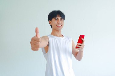 Kameraya gülümseyen mutlu Güneydoğu Asyalı adam akıllı telefonu tutarken başparmağını kaldırıyor. Beyaz arkaplan ile izole edilmiş portre, onaylama ve pozitifliği ifade ediyor