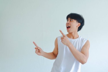 Spor kıyafetli heyecanlı genç Asyalı adam beyaz arka planda izole edilmiş boş bir yeri işaret ediyor. Reklamcılık, fitness promosyonu ve sağlık yaşam tarzı içeriği için mükemmel