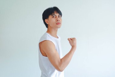 Jimnastik salonundaki kendine güvenen genç Asyalı adam kollarını esnetip ciddi bir ifade takınıyor, kameraya güç ve güç gösteriyor, beyaz arka planda izole edilmiş, fitness ve egzersiz konsepti.