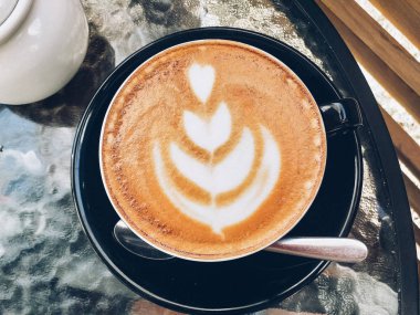 Sütlü kahvenin en üst görüntüsü, sütlü siyah bir fincanda latte sanatı ve cam bir masanın üzerinde köpük desenli fincan tabağı. Resim dizme yöntemleri.
