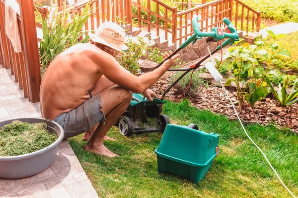 Doğal arka plan. Yazın bir çiçek bahçesinin yakınındaki kendi bahçesinden çim biçme makinesini temizleyen bir adam. Yan görüş, yatay. Tasarım, doğa ve peyzaj kavramı.