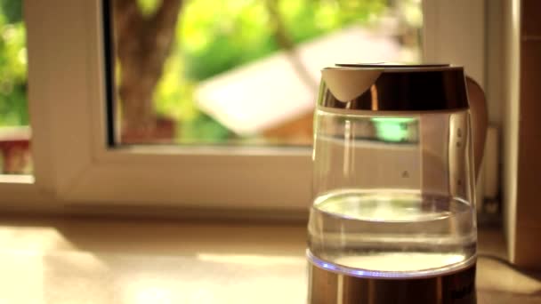 Een transparante theepot met water kookt tegen een zonsondergang die door een raam schijnt. Het concept van een koffiepauze en het einde van de dag. Warme zomeravond ontspanning. — Stockvideo