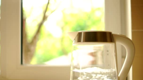 Une théière transparente avec de l'eau se trouve sur la table de cuisine. le processus d'ébullition de l'eau contre un coucher de soleil qui brille à travers une fenêtre. Le concept d'une pause café et la fin de la journée. — Video