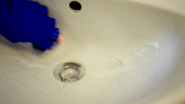 Kvinnliga händer i blå latexhandskar gnider handfatet med en tvättlapp. Det är mycket skum runt handfatet. Konceptet att hålla badrummet rent. — Stockvideo