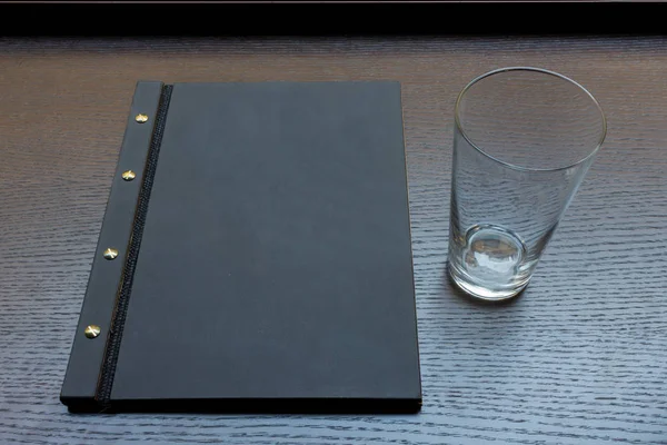 Black or dark grey restaurant menu with copy space and empty glass on dark wooden table background