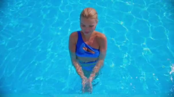 Дівчина в блакитному купальнику стоїть в басейні і грає з водою для камери, блакитні брижі води, що світяться на сонці .. — стокове відео