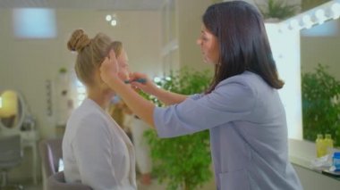 Makyajcı bir kız sarışına makyaj yapar, iki kız makyaj yapar, profesyonel bir makyaj sanatçısı, esmer bir kadının profesyonel makyajı.