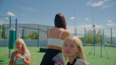 Kadınlar şehirdeki spor sahasında antrenman yapıyor, genç sarışın kameralar önünde çömelmiş, günlük spor tarzında.