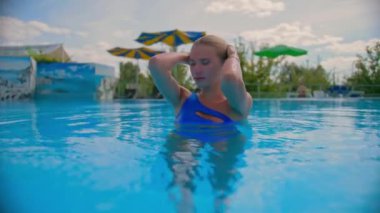 Kar beyazı gülüşlü bir kız suyun içinde duruyor, mutlu bir sarışın kameraya gülümsüyor ve havuzda yüzüyor..