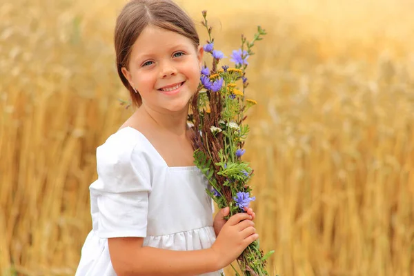 Klein meisje met een boeket wilde bloemen in haar handen in een tarweveld. — Stockfoto