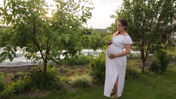 En gravid mamma går i trädgården nära huset. — Stockvideo