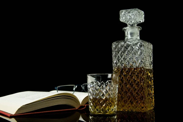 Băutură alcoolică în decantor de cristal. Într-un pahar turnat whisky pe sticlă neagră și fundal negru. Deschide o carte cu ochelari . — Fotografie, imagine de stoc