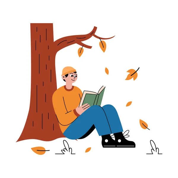 Çocuk ağacın altında oturuyor ve sonbahar parkında kitap okuyor.