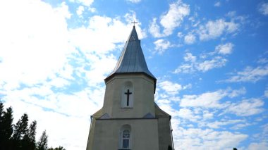 Bir kilise kulesi, tüylü beyaz bulutlarla süslenmiş parlak mavi gökyüzünü deler. Güneş ışığında yıkanan bir çan kulesi dimdik duruyor. Geniş alana karşı bir haç silueti..