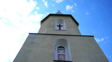 Bir kilise kulesi, tüylü beyaz bulutlarla süslenmiş parlak mavi gökyüzünü deler. Güneş ışığında yıkanan bir çan kulesi dimdik duruyor. Geniş alana karşı bir haç silueti..