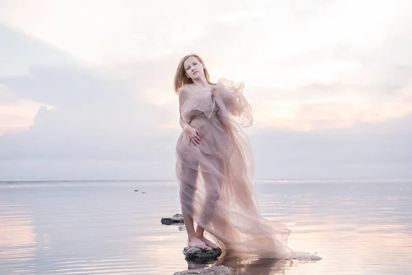 A girl in a weightless light fabric of pastel tones in the water of a calm pond with a reflection of the sunset gentle sky in the water. Artistic image of a gentle girl in nature.