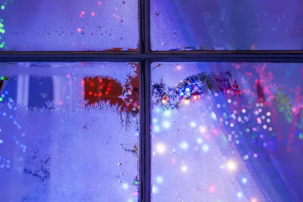 The night window with frosty patterns and bright multicolored Christmas. New Year\'s lights and decorations outside the window.