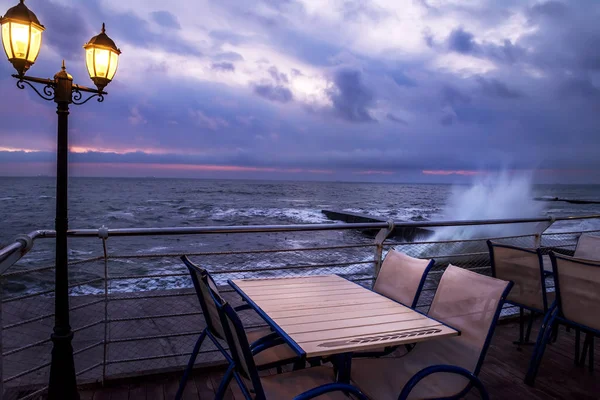 Dramatic sunrise by the sea. Cafe tables on the pier and the waves beating on the pier. Dark night photo.