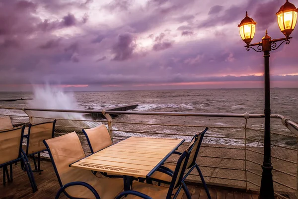 Dramatic sunrise by the sea. Cafe tables on the pier and the waves beating on the pier. Dark night photo.