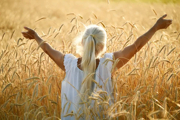 An old woman with long gray hair stands with her back with her arms spread out to the side in a field with golden ears of cereal crops in the sunset light.