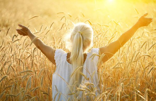 An old woman with long gray hair stands with her back with her arms spread out to the side in a field with golden ears of cereal crops in the sunset light.