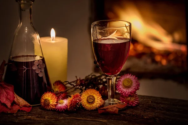 Cozy evening still life by . A glass of red wine, an old decanter, green leaves and dried flowers against a burning fire. Dark photo. The cozy atmosphere of an autumn, winter evening.