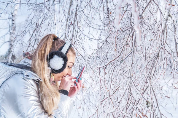 The girl enthusiastically looks at the screen of her phone in the winter park. Tree branches in the park covered with magical sparkling ice and snow.