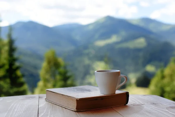 A coffee cup and a book on a wooden table against the backdrop of a beautiful green mountain landscape. Relax outdoor recreation.