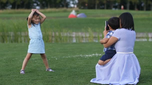 Мама фотографирует смешную азиатскую девочку в парке — стоковое видео